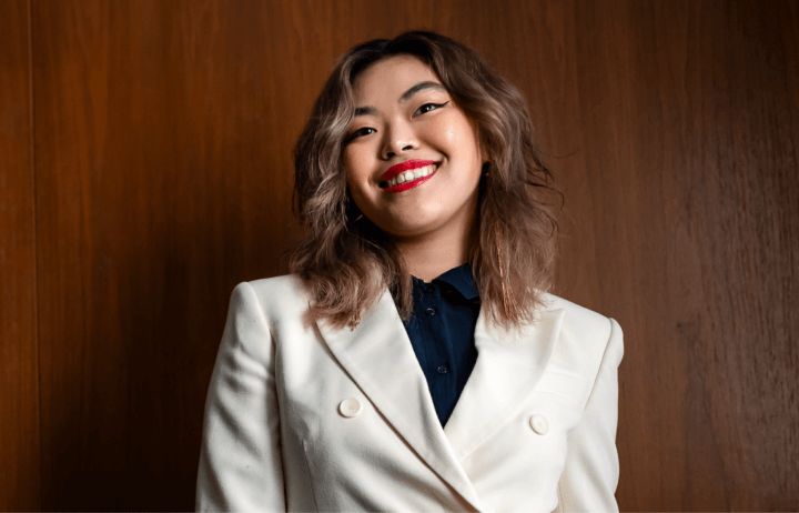 Young person with long brunette hair wearing a white suit smiling against a wood backdrop