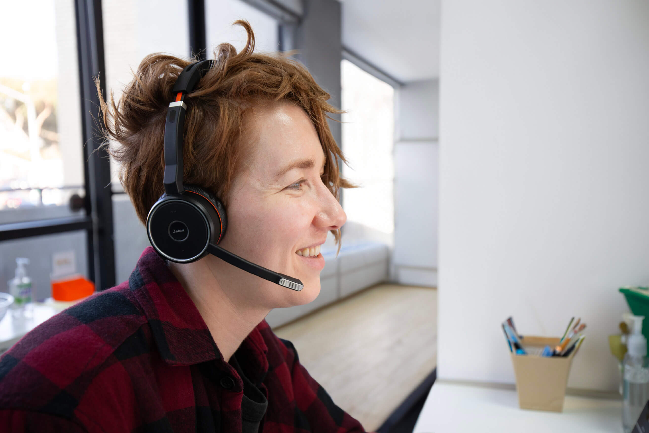 A young adult wearing a headset, sits at a desk in a brightly lit room