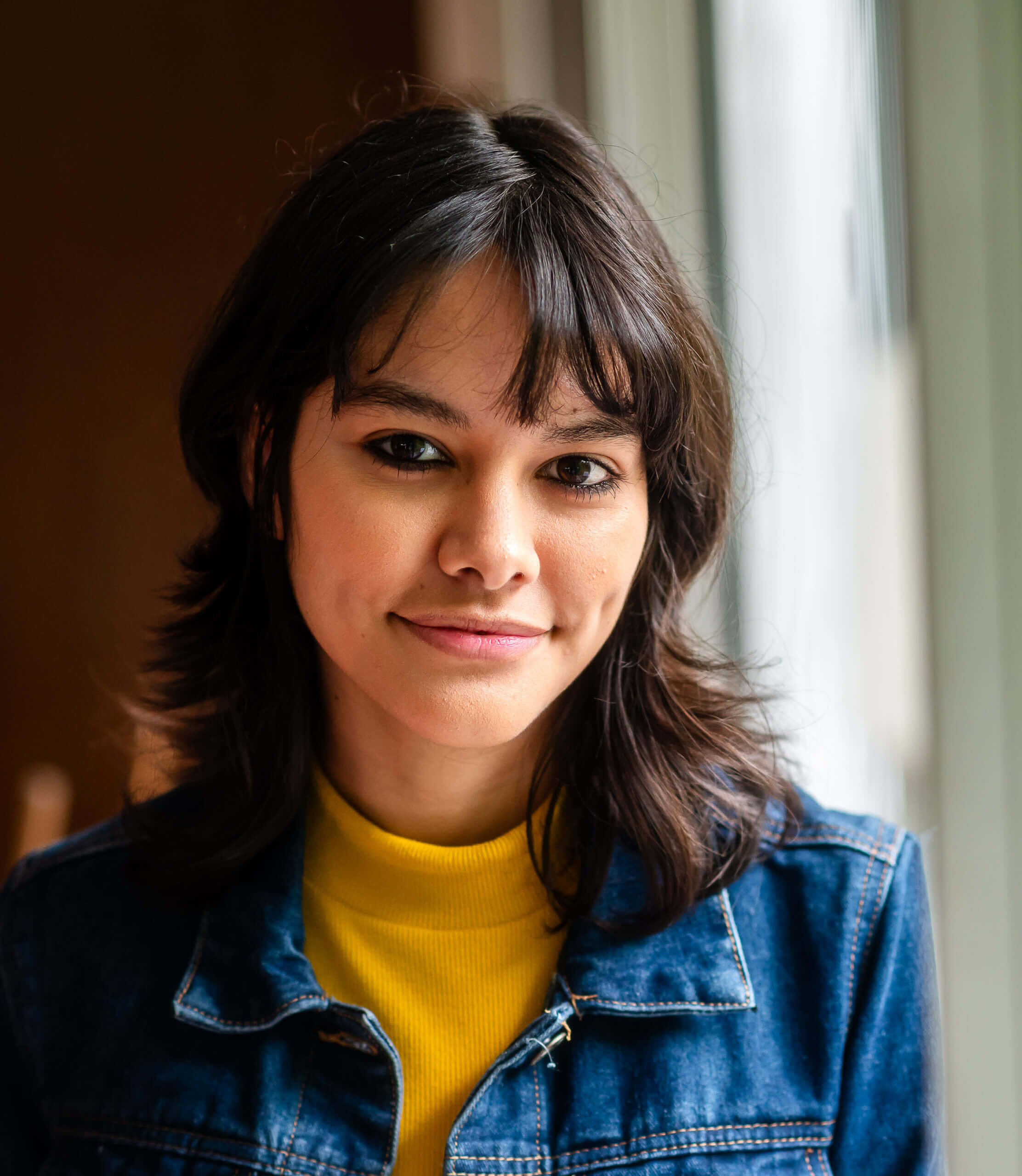 An LGBTQ+ young person leans against a window in a jean jacket.