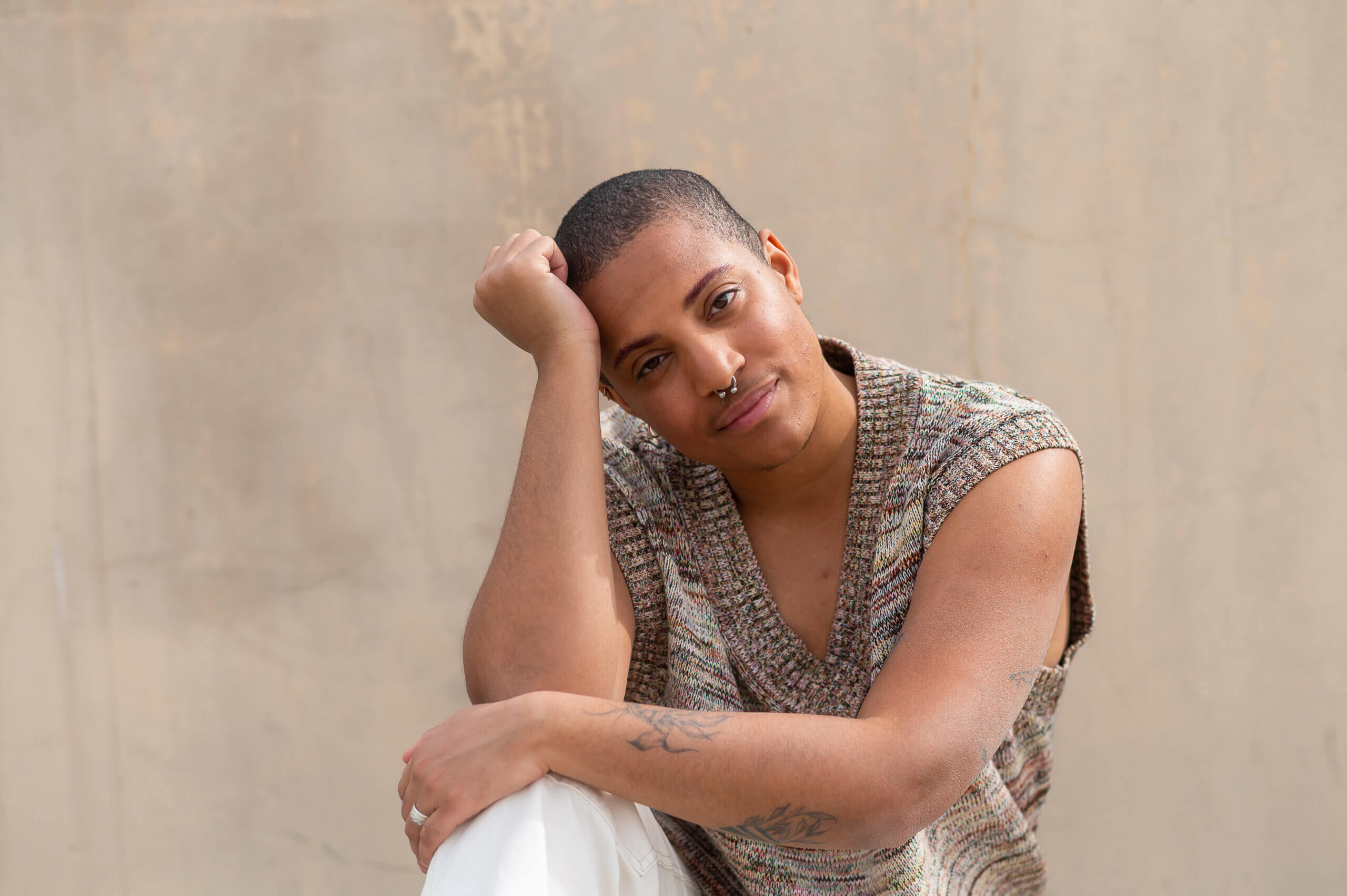An LGBTQ+ young person with a nose ring smiles in front of a plain background.