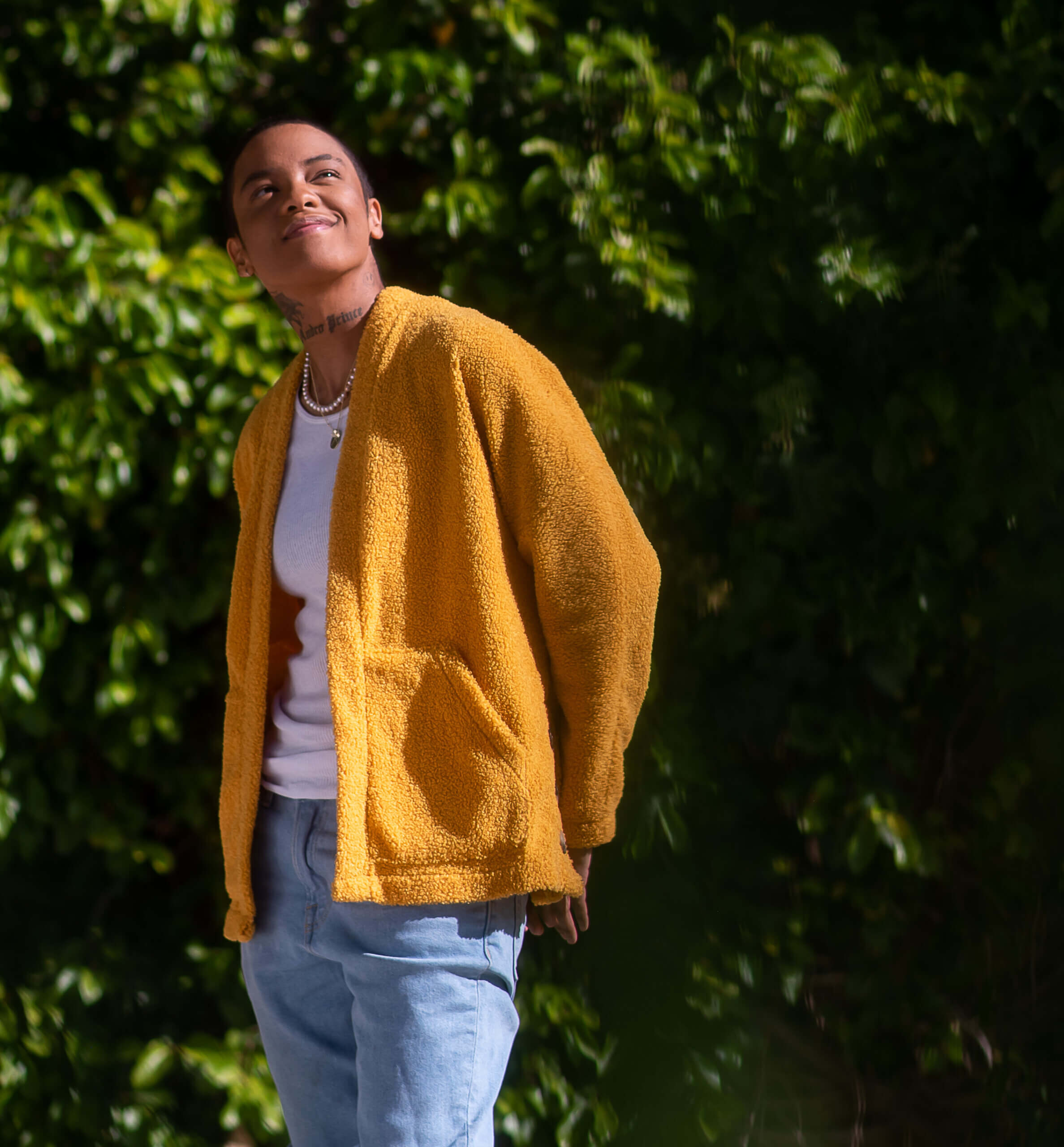 An LGBTQ+ young person smiles and looks up in front of foliage.